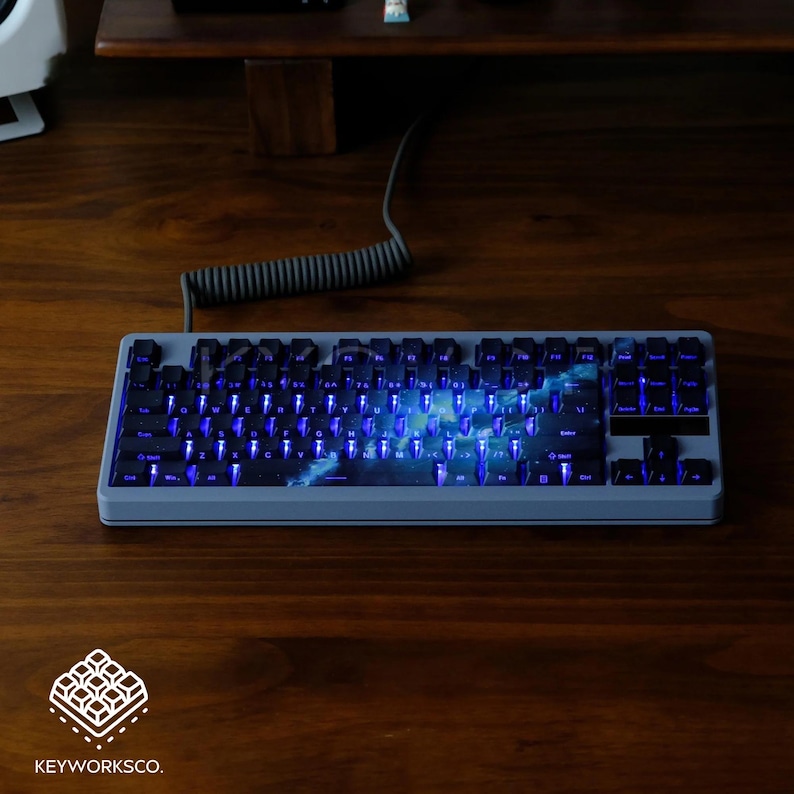 May include: A gray mechanical keyboard with blue backlighting and a space-themed design. The keyboard is on a wooden surface, with a coiled cable and the Keyworks Co. logo visible.
