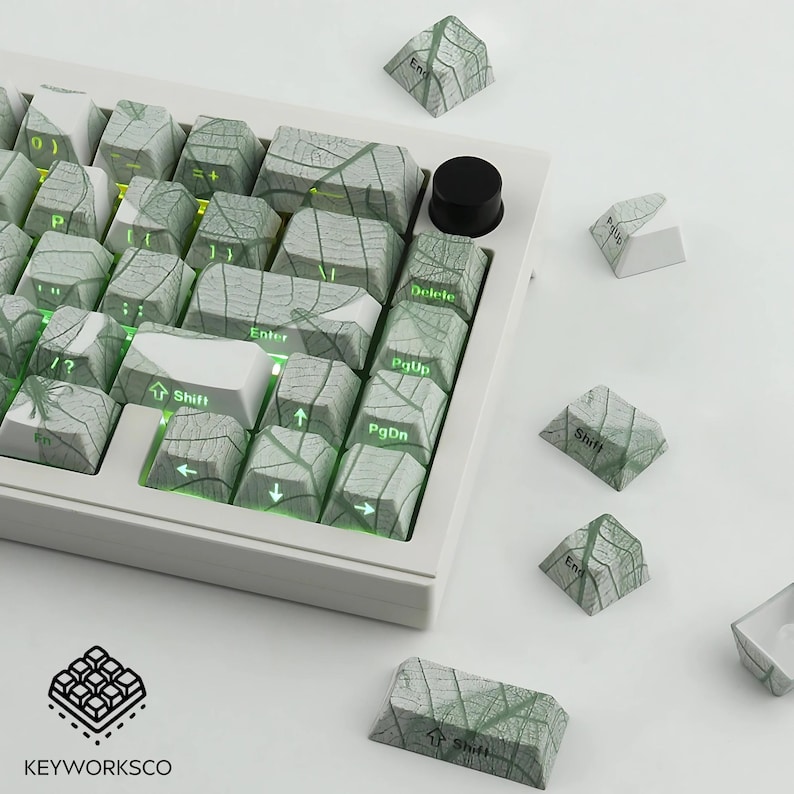 K&ouml;nnte beinhalten: Eine wei&szlig;e mechanische Tastatur mit individuellen Tastenkappen mit einem Blattdesign in Grau- und Gr&uuml;nt&ouml;nen. Die Tastatur hat einen schwarzen Lautst&auml;rkeregler und mehrere lose Tastenkappen. Das Markenlogo "KEYWORKSCO" ist unten links sichtbar.