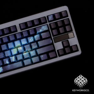 May include: A mechanical keyboard with a light gray frame and dark gray keycaps. The keycaps feature a blue and purple galaxy design. The keyboard is angled towards the viewer, with the KeyworksCo. logo in the bottom right corner.