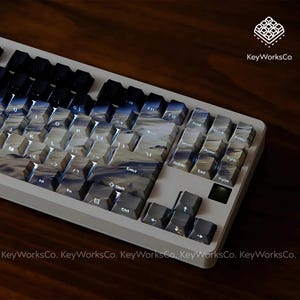 May include: A mechanical keyboard with custom keycaps. The keycaps have a gradient design in blue, grey, and white, like a cloudy sky. The keyboard has a white frame and black keys. The brand name "KeyWorksCo" is visible.