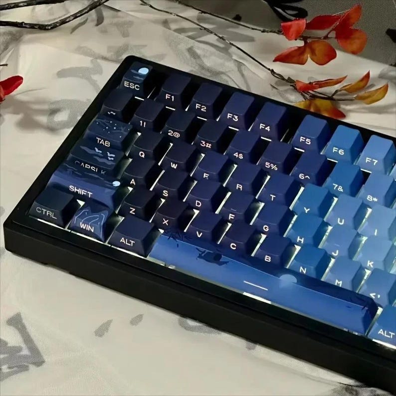 May include: A blue and black mechanical keyboard with keycaps featuring a gradient of blue shades and white text. The keyboard has a black frame and a space bar with a moon design.