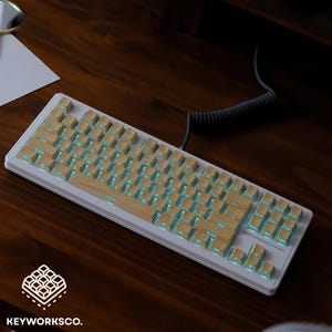 Puede incluir: Un teclado mecánico blanco con teclas de madera y retroiluminación azul. El teclado está sobre una superficie de madera oscura. El logotipo de KEYWORKSCO. está en la esquina inferior izquierda.