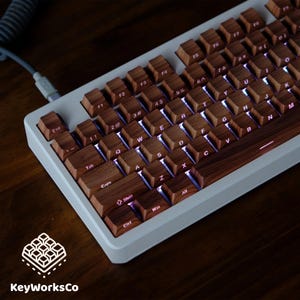 May include: A mechanical keyboard with wooden keycaps and a white frame. The keys are illuminated with a soft, purple glow. The keyboard is connected to a gray coiled cable. The KeyWorksCo logo is in the lower left corner.