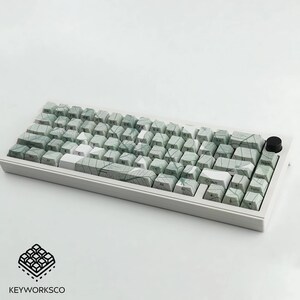 K&ouml;nnte beinhalten: Eine wei&szlig;e mechanische Tastatur mit gr&uuml;nen und wei&szlig;en Tasten. Die Tasten haben ein blattartiges Muster. Die Tastatur hat einen schwarzen Knopf oben rechts. Das Logo "KEYWORKSCO" befindet sich unten links.