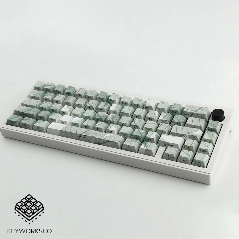 K&ouml;nnte beinhalten: Eine wei&szlig;e mechanische Tastatur mit gr&uuml;nen und wei&szlig;en Tasten. Die Tasten haben ein blattartiges Muster. Die Tastatur hat einen schwarzen Knopf oben rechts. Das Logo "KEYWORKSCO" befindet sich unten links.