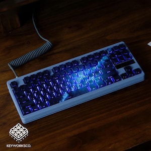 May include: A gray mechanical keyboard with backlit keys, displaying a galaxy design. The keyboard sits on a wooden surface, with a coiled cable visible. The keys are black with blue backlighting, and the keyboard has a rectangular shape.