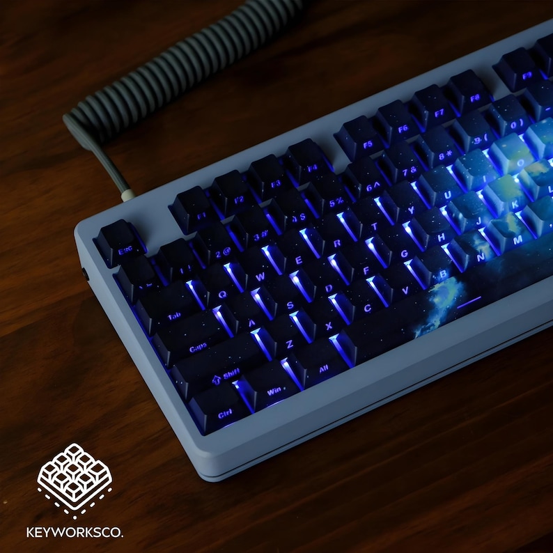 May include: A custom mechanical keyboard with black keycaps and blue backlighting. The keyboard has a light blue frame and a space-themed design on the base. A coiled gray cable is connected to the keyboard. The logo "KEYWORKSCO." is visible.