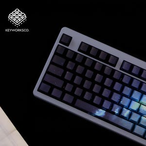 May include: A gray mechanical keyboard with dark keycaps. The keycaps have a starry night design with blue and white accents. The keyboard is angled, and the logo "KEYWORKSCO." is in the upper left corner.