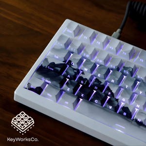 Puede incluir: Teclado mecánico blanco con un diseño de paisaje en las teclas. Las teclas están retroiluminadas con un brillo púrpura. El teclado tiene un cable en espiral y el logotipo "KeyWorksCo." es visible.