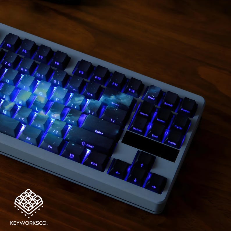 May include: A backlit mechanical keyboard with a white frame and black keys. The keycaps feature a galaxy design with blue and white hues. The keyboard has a full layout with function keys and a number pad. The brand logo "KEYWORKSCO." is visible.
