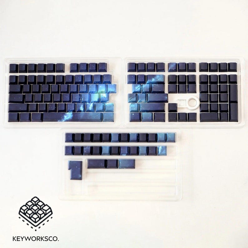 May include: Keyboard keycaps in clear plastic trays. The keycaps are primarily dark blue with a space-themed design, featuring shades of blue and white. The logo "KEYWORKSCO." is visible in the bottom left corner.
