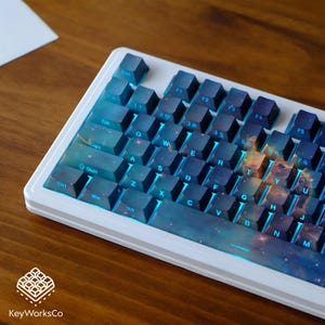 Puede incluir: Un teclado mecánico blanco con teclas azules y con temática de galaxia. El teclado tiene un marco blanco y retroiluminación azul. Las teclas son un degradado de azul y presentan un diseño de galaxia. El teclado tiene un diseño QWERTY estándar.