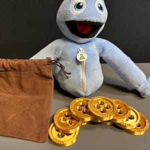 May include: A blue plush toy with a zipper and a green and white emblem, a brown drawstring bag, and several gold-colored coins with a three-hole design. The coins are stacked in a pile.