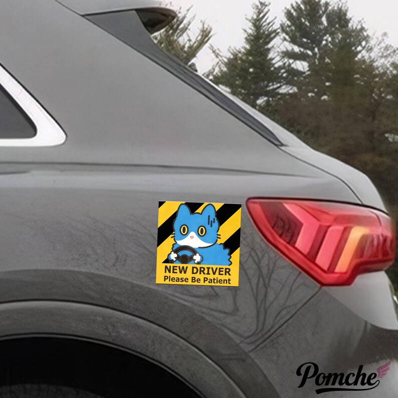 May include: A square sticker on a car showing a blue cat driving. The sticker has a yellow and black striped background with the text "NEW DRIVER Please Be Patient". The car is grey.