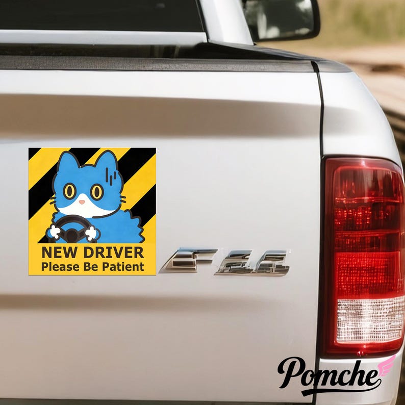 May include: A square sticker on a white vehicle shows a blue cartoon cat driving. The cat has large, round eyes and holds a steering wheel. The sticker has a yellow and black striped background and the text "NEW DRIVER Please Be Patient".