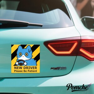 May include: A car magnet featuring a blue cat driving, set against a yellow and black striped background. The text "NEW DRIVER Please Be Patient" is below the cat. The magnet is attached to the rear of a light blue car.