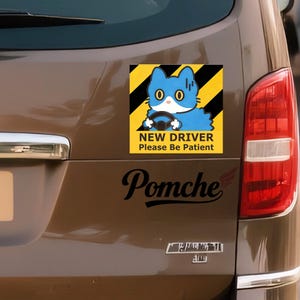May include: A brown car with a sticker that says "NEW DRIVER Please Be Patient". The sticker features a blue cat driving a car on a yellow and black background. The word "Pomche" is written in black script below the sticker.