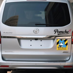 May include: The rear of a silver vehicle with a "Pomche" decal and a "New Driver Please Be Patient" sticker. The sticker shows a blue cat and has a yellow and black striped background.