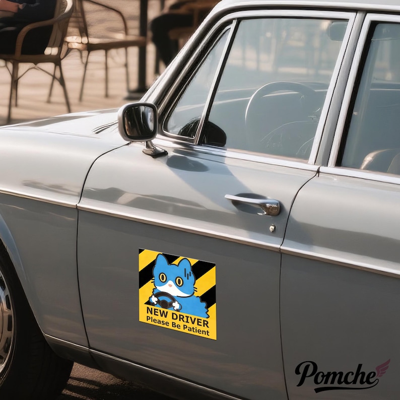May include: A rectangular car magnet showing a blue cartoon cat driving a car, with the text "NEW DRIVER Please Be Patient" on a yellow and black striped background. The magnet is attached to the side of a light grey vintage car.