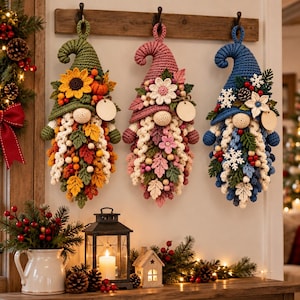 May include: Three hanging crochet gnome ornaments featuring seasonal themes. The left gnome has a green hat with sunflowers and autumn leaves. The center gnome has a pink hat with flowers. The right gnome has a blue hat with snowflakes and pinecones.