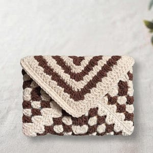May include: A rectangular envelope-style clutch bag crafted from crocheted yarn. The accessory features a repeating geometric pattern in alternating shades of dark brown and cream, with a triangular flap closure that secures the main compartment.