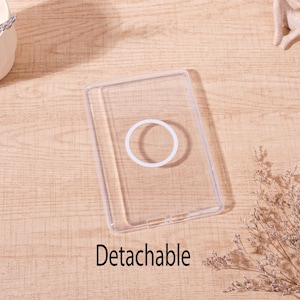 Op de afbeelding: Een transparante, rechthoekige telefoonhoes met een witte, cirkelvormige grip. Het woord "Detachable" staat in het zwart onderaan gedrukt. De hoes ligt op een lichtbruin houten oppervlak.