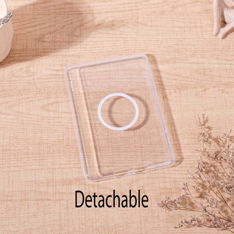 Op de afbeelding: Een transparante, rechthoekige telefoonhoes met een witte, cirkelvormige grip. Het woord "Detachable" staat in het zwart onderaan gedrukt. De hoes ligt op een lichtbruin houten oppervlak.