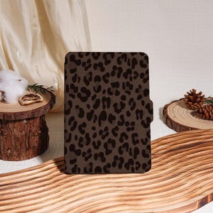 Puede incluir: Una funda de tableta marrón con un estampado de leopardo marrón oscuro. La funda rectangular tiene un cierre magnético. Se muestra sobre una superficie de madera.