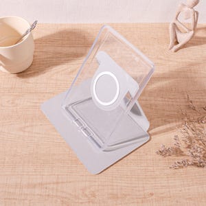 Op de afbeelding: Een doorzichtige plastic telefoonstandaard met een witte, cirkelvormige magnetische bevestiging. De standaard is schuin en bevestigd aan een lichtgrijze basis. De standaard staat op een houten oppervlak.