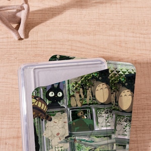 Op de afbeelding: Een doorzichtige plastic container met een deksel, met een kleurrijk grafisch ontwerp. Het ontwerp bevat stripfiguren, waaronder een zwarte kat met grote ogen en andere grillige figuren. De container staat tegen een lichte houten achtergrond.