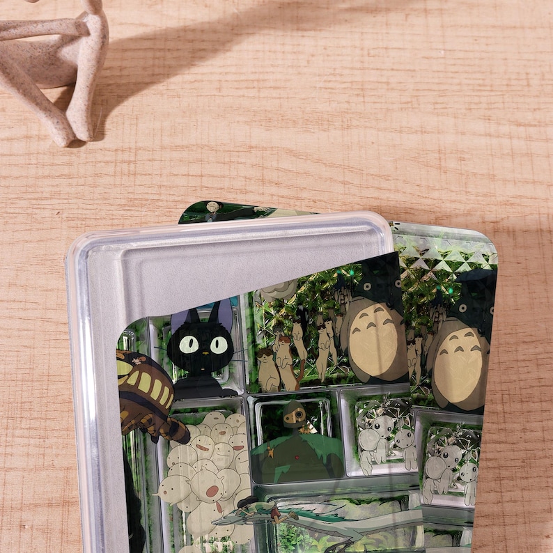 Op de afbeelding: Een doorzichtige plastic container met een deksel, met een kleurrijk grafisch ontwerp. Het ontwerp bevat stripfiguren, waaronder een zwarte kat met grote ogen en andere grillige figuren. De container staat tegen een lichte houten achtergrond.