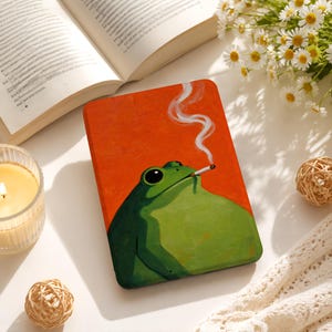 Può includere: Una custodia rettangolare per e-reader con un'illustrazione in stile pittura a olio di una rana verde con grandi occhi neri che fuma una sigaretta, su uno sfondo arancione vibrante con una sottile scia di fumo bianco che sale dalla sigaretta.