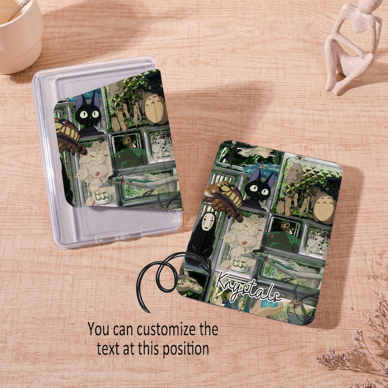 Op de afbeelding: Twee doorzichtige telefoonhoesjes met een kleurrijk ontwerp in anime-stijl met personages als Jiji de kat en Totoro. E&eacute;n hoesje is open en de andere is gesloten. De tekst "Krystals" is zichtbaar op &eacute;&eacute;n hoesje en de tekst "You can customize the text at this position" is zichtbaar op de andere.