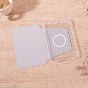 Op de afbeelding: Een lichtgrijze tablethoes met een transparant plastic frame. De hoes heeft een cirkelvormige uitsparing met een witte ring. De hoes is open en onthult de binnenkant. De hoes is ontworpen om een tablet te beschermen.