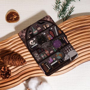 May include: A dark gray decorative mat featuring a whimsical illustration of a witch's bookshelf. The design includes potion bottles, a skull, a cat, books, and a spiderweb. The mat is placed on a wooden surface with decorative elements.