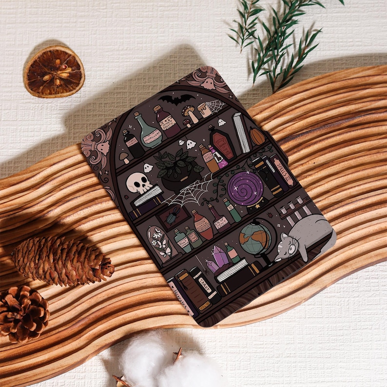 May include: A dark gray decorative mat featuring a whimsical illustration of a witch's bookshelf. The design includes potion bottles, a skull, a cat, books, and a spiderweb. The mat is placed on a wooden surface with decorative elements.
