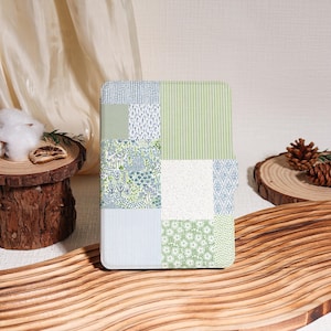 May include: A square, patchwork-style case featuring various patterns in shades of green, blue, and white. The patterns include stripes, floral designs, and dotted textures. The case is displayed on a wooden surface.