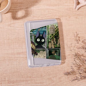 Op de afbeelding: Een doorzichtige plastic doos met een scharnierend deksel, met een kleurrijke collage van illustraties in anime-stijl. De afbeeldingen bevatten een zwarte kat met grote ogen, een bruin insect en andere stripfiguren. De doos staat op een lichtbruin houten oppervlak.