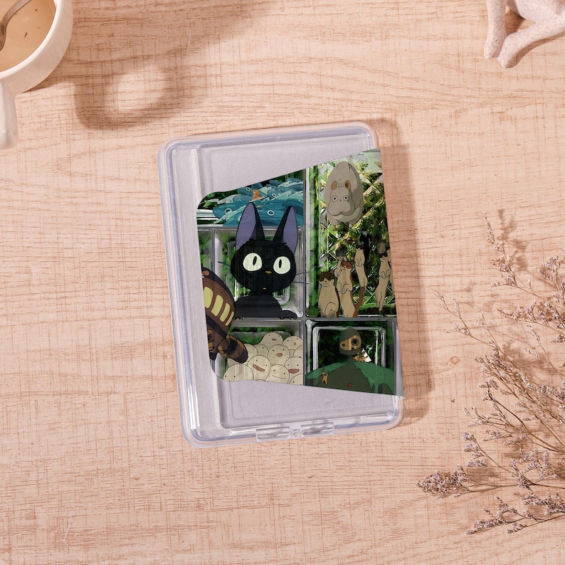 Op de afbeelding: Een doorzichtige plastic doos met een scharnierend deksel, met een kleurrijke collage van illustraties in anime-stijl. De afbeeldingen bevatten een zwarte kat met grote ogen, een bruin insect en andere stripfiguren. De doos staat op een lichtbruin houten oppervlak.
