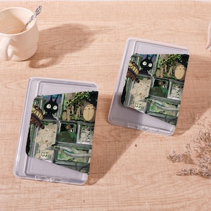Op de afbeelding: Twee doorzichtige plastic telefoonhoesjes met een kleurrijk grafisch ontwerp met personages uit een animatiefilm. De hoesjes liggen op een lichtbruin houten oppervlak, met een witte mok en een lepel op de achtergrond.