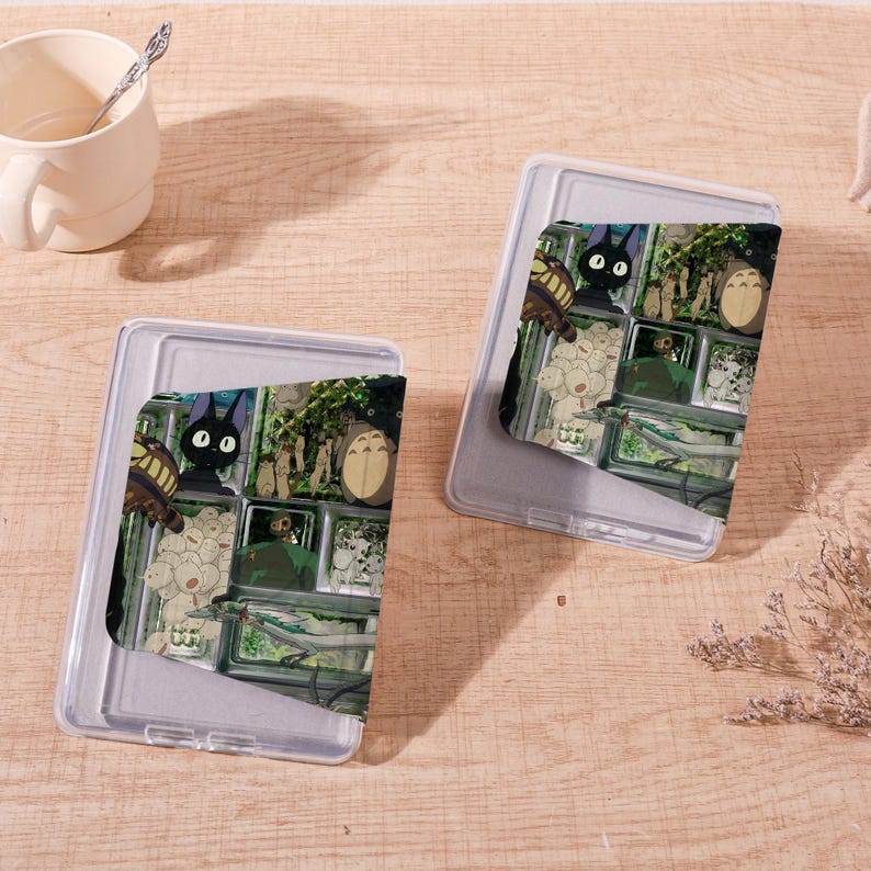 Op de afbeelding: Twee doorzichtige plastic telefoonhoesjes met een kleurrijk grafisch ontwerp met personages uit een animatiefilm. De hoesjes liggen op een lichtbruin houten oppervlak, met een witte mok en een lepel op de achtergrond.