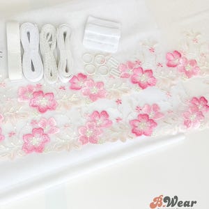 Könnte beinhalten: Ein weißer Stoff mit rosa floraler Spitzenborte, weißen elastischen Trägern, weißen BH-Verschlüssen und weißen Ringen. Der Stoff ist für die Herstellung eines BHs bestimmt.