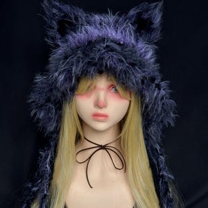 May include: A faux fur hat with pointed ears, in shades of purple and black, sits atop a mannequin head with long blonde hair. The hat has a fluffy texture and is styled to resemble a wolf or cat. A dark brown ribbon is tied around the neck.