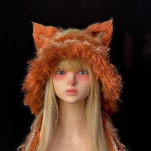 Op de afbeelding: Een pop met een harige oranje vossenhoed met puntoren. De hoed heeft een ruige textuur en is afgezet met witte en oranje strengen. De pop heeft lang blond haar, tegen een zwarte achtergrond.