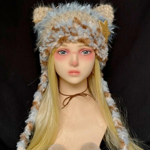 Puede incluir: Un gorro afelpado con orejas de gato en tonos beige, azul y marrón, adornado con una estrella dorada. El gorro tiene largas solapas para las orejas y dos pequeños pompones beige. Lo lleva un maniquí con pelo rubio y una gargantilla marrón.