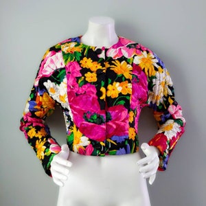 Puede incluir: Una chaqueta acolchada negra con estampado floral con flores rosas, amarillas y blancas. La chaqueta tiene cierre de botón y mangas largas.