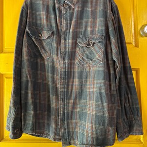 May include: A long-sleeved, button-down flannel shirt in a plaid pattern of blue, brown, and cream. The shirt has two chest pockets with button closures and a classic collar. The shirt is hanging on a black hanger.