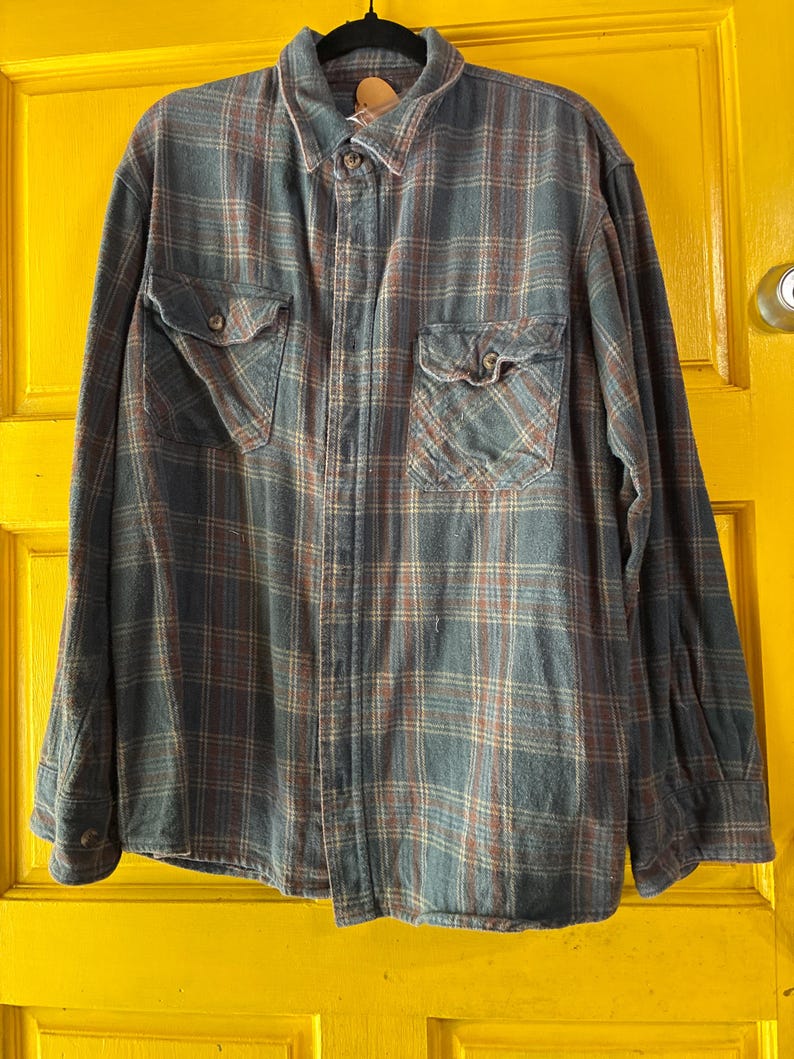 May include: A long-sleeved, button-down flannel shirt in a plaid pattern of blue, brown, and cream. The shirt has two chest pockets with button closures and a classic collar. The shirt is hanging on a black hanger.