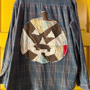 May include: Blue plaid flannel shirt featuring a patchwork pumpkin design on the back. The pumpkin is crafted from diverse fabric scraps in brown, white, and colourful patterns, with triangular eyes and a stitched mouth.