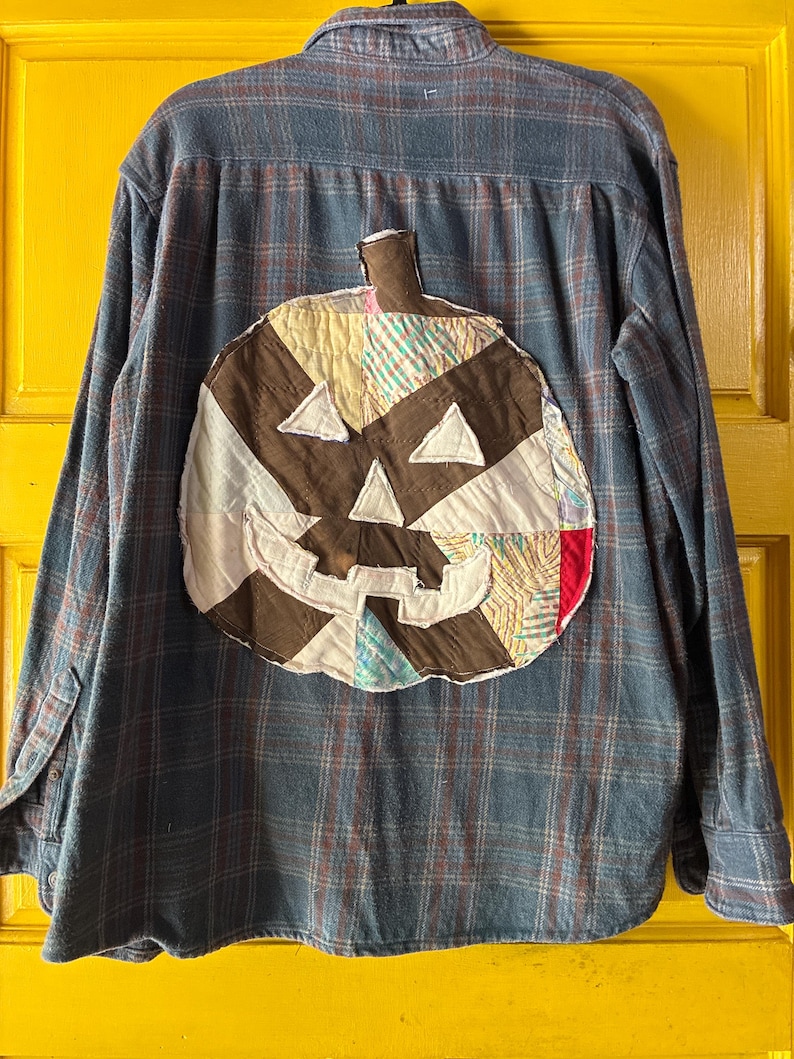 May include: Blue plaid flannel shirt featuring a patchwork pumpkin design on the back. The pumpkin is crafted from diverse fabric scraps in brown, white, and colourful patterns, with triangular eyes and a stitched mouth.